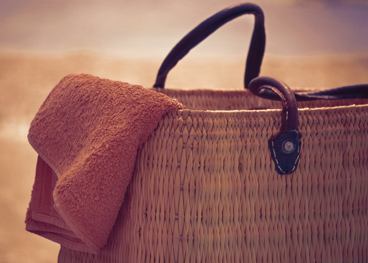 A woven straw beach bag with leather handles and a coral folded towel in warm afternoon light